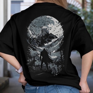 Op de afbeelding: Zwart T-shirt met een gedetailleerde illustratie van een fantasie scène. Het ontwerp bevat een silhouet met zwaarden, een wolf en een berglandschap met een volle maan en vogels. Het kunstwerk is in grijstinten en wit.