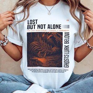Può includere: Una t-shirt bianca con una stampa grafica scura di una creatura scheletrica e un esploratore con una torcia. Il testo recita LOST BUT NOT ALONE, THE LOST RIVER DOESN'T FORGIVE, IT REMEMBERS, GHOSTS LURK BELOW e altro testo descrittivo.
