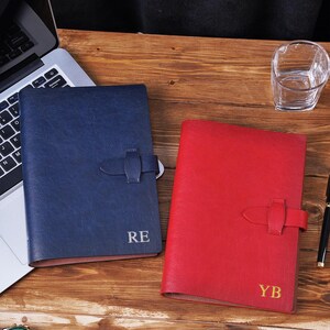 May include: A navy blue notebook with the initials RE and a red notebook with the initials YB, both featuring a textured finish, a vertical strap closure, and a rectangular shape. These two notebooks are positioned side by side on a wooden surface.