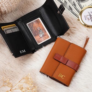 May include: Two wallets, one black and one brown, are displayed on a fluffy white surface. The black wallet is open, revealing cards and a photo. The brown wallet is closed with a brown strap. Both wallets have initials.