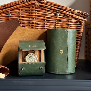 Könnte beinhalten: Ein grünes Leder-Uhrenetui und eine zylindrische Uhrenrolle werden gezeigt. Das mit "P.G." geprägte Etui enthält eine goldfarbene Uhr. Die mit "SJ" markierte Rolle steht daneben. Ein Weidenkorb und Lederstücke sind im Hintergrund.