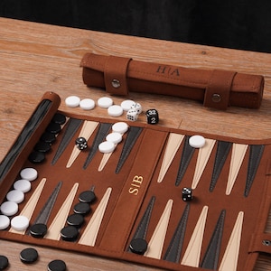 May include: A brown leather backgammon set with black and white game pieces. The set is open, displaying the playing surface with black, white, and tan triangles. A brown leather dice cup and a roll-up case with initials are also visible.
