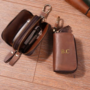 May include: Two brown leather key cases are shown. One is open, displaying keys and a phone, the other is closed with the initials "B.C." in gold. Both have zips and metal clasps. A brown and green leather notebook is in the background.