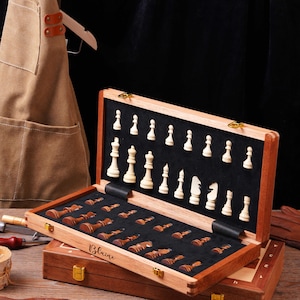 May include: A wooden chess set featuring a hinged storage box with a black interior lining. The set includes light-colored and dark-colored wooden chess pieces held in custom-fitted slots. The name Blaine is engraved on the side of the box.