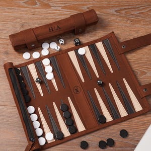 A leather roll-up backgammon set shown open with playing pieces arranged across the board. The exterior strap with personalised initials sits above the board, demonstrating custom engraving and the portable travel-friendly format.