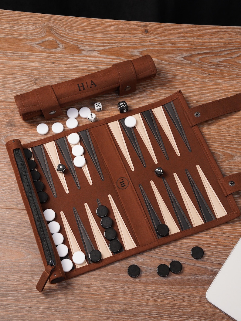 A leather roll-up backgammon set shown open with playing pieces arranged across the board. The exterior strap with personalised initials sits above the board, demonstrating custom engraving and the portable travel-friendly format.