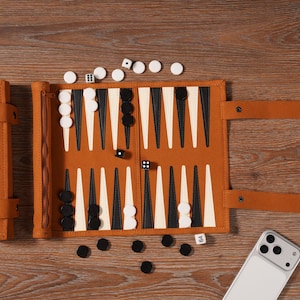 May include: A tan leather backgammon set with black and white game pieces, dice, and a matching leather case. The case is embossed with the initials "BLD". The set is open on a wooden surface.