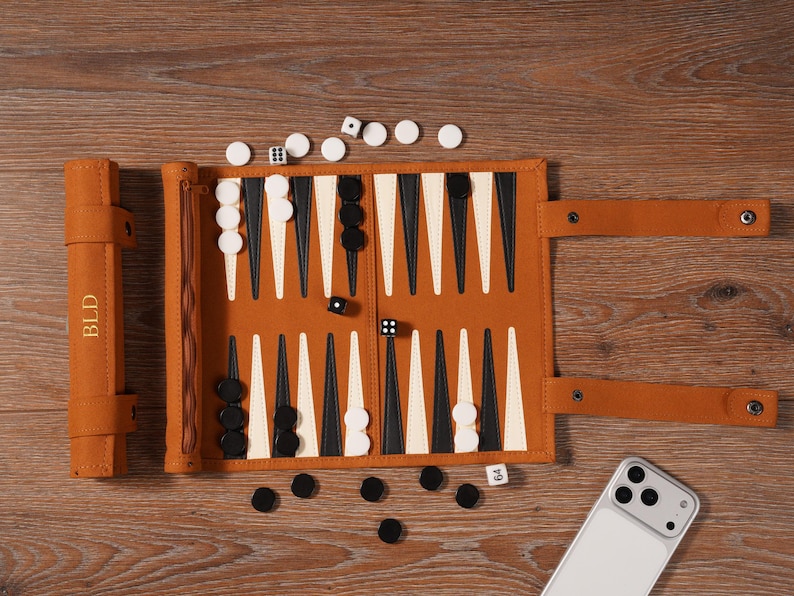 May include: A tan leather backgammon set with black and white game pieces, dice, and a matching leather case. The case is embossed with the initials "BLD". The set is open on a wooden surface.