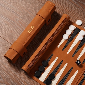 May include: A brown backgammon set with a zippered case and a roll-up case. The backgammon board features black and white triangles. The roll-up case has the initials "BLD" in gold lettering. Black and white game pieces and a die are visible.