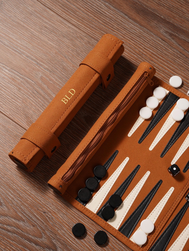 May include: A brown backgammon set with a zippered case and a roll-up case. The backgammon board features black and white triangles. The roll-up case has the initials "BLD" in gold lettering. Black and white game pieces and a die are visible.