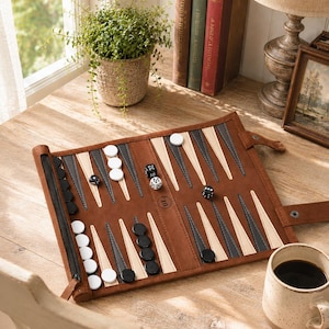 May include: A brown leather backgammon set with black, white, and grey game pieces. The board is open, with dice scattered across the playing surface. A cup of coffee and a small potted plant are also visible.