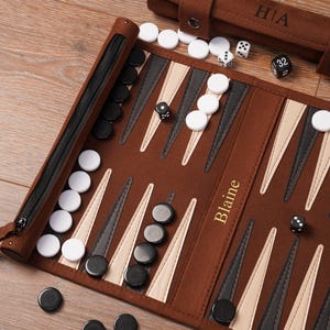 A personalised leather backgammon board with the name “Blaine” engraved in gold foil. Black and white checkers and dice are scattered across the playing surface, highlighting craftsmanship, customisation, and the roll-up travel design.