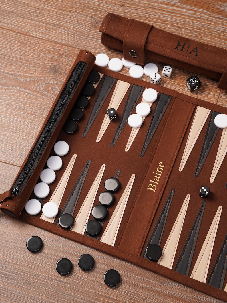 A personalised leather backgammon board with the name “Blaine” engraved in gold foil. Black and white checkers and dice are scattered across the playing surface, highlighting craftsmanship, customisation, and the roll-up travel design.