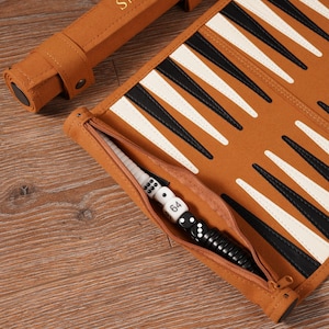 May include: A brown backgammon set with black and white triangles. The set includes a zippered pouch with dice and a dice cup. The rolled-up case has the initials "SIB" embossed on it.