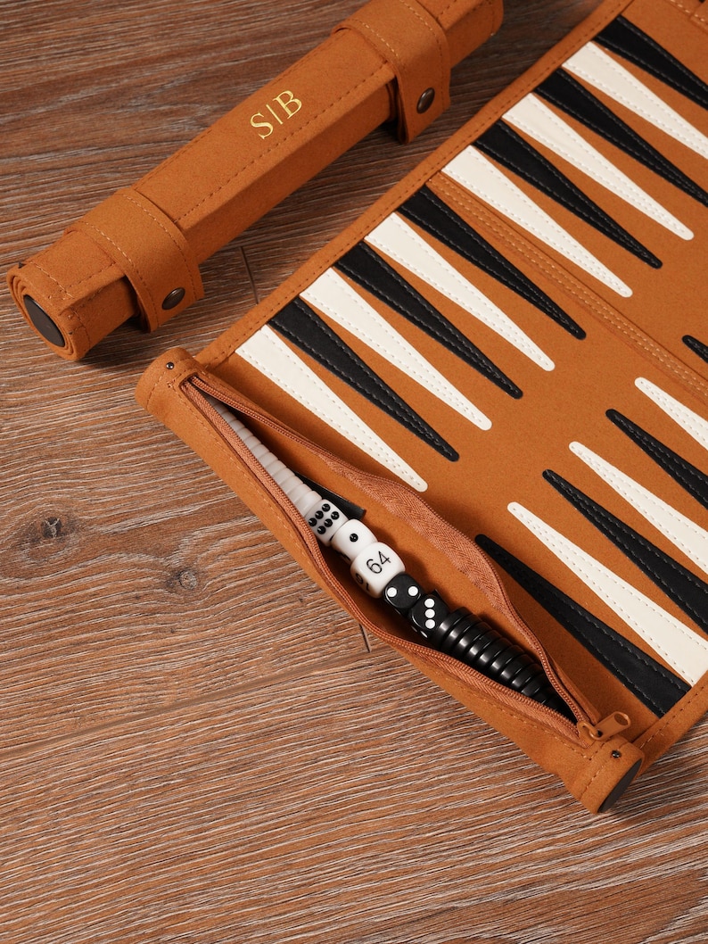 May include: A brown backgammon set with black and white triangles. The set includes a zippered pouch with dice and a dice cup. The rolled-up case has the initials "SIB" embossed on it.