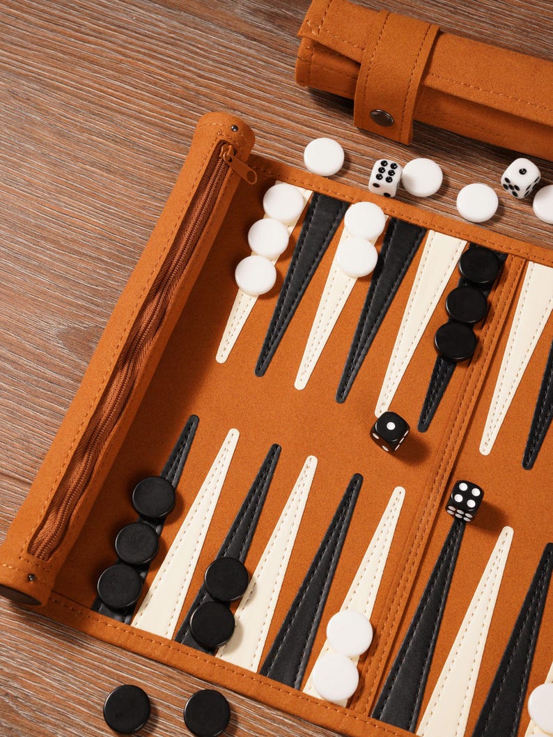 May include: A brown backgammon set with white and black game pieces. The board is open, showing the playing surface with black and white triangles. Dice and a zippered case are also visible. The set is on a wooden surface.