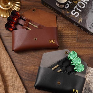 May include: Two envelope-style dart cases made of leather, one brown with the initials FC and one black with the initials PB. Each case features a metal snap closure and holds three darts with distinct flight patterns, including red flame and green target designs.