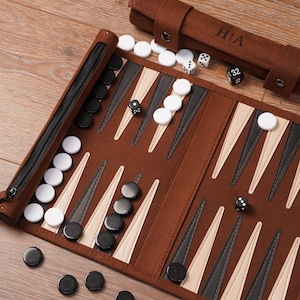 May include: A brown backgammon set with black and white counters, dice, and a zippered case. The game board is open, showing the triangular points and the arrangement of the pieces. A brown roll-up case with the initials "HIA" is also visible.