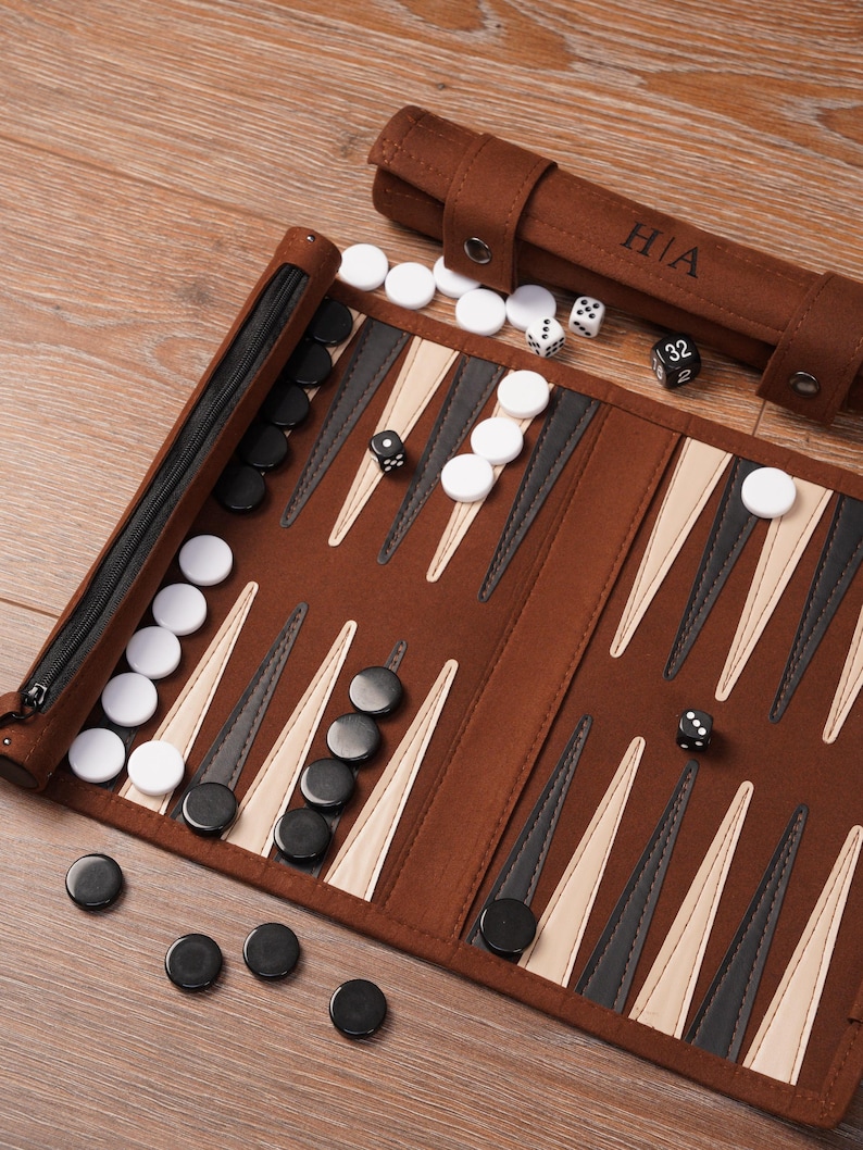 May include: A brown backgammon set with black and white counters, dice, and a zippered case. The game board is open, showing the triangular points and the arrangement of the pieces. A brown roll-up case with the initials "HIA" is also visible.