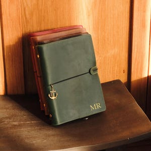 May include: A stack of leather journals in shades of green, brown, and pink. The front journal is dark green with gold-colored initials "MR" and an anchor charm. The journals are closed with an elastic band.