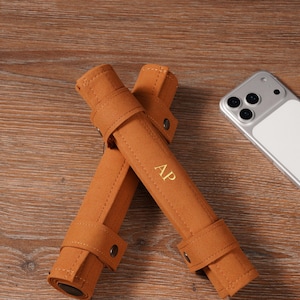 May include: Two brown, rolled felt pen cases with snap closures, each featuring the gold initials "AP". A smartphone with a white back is visible in the background. The cases are on a wooden surface.