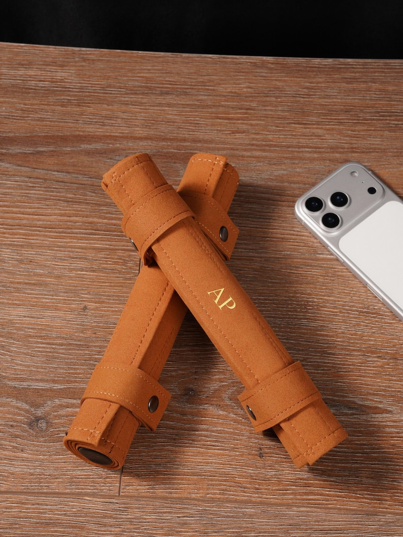 May include: Two brown, rolled felt pen cases with snap closures, each featuring the gold initials "AP". A smartphone with a white back is visible in the background. The cases are on a wooden surface.