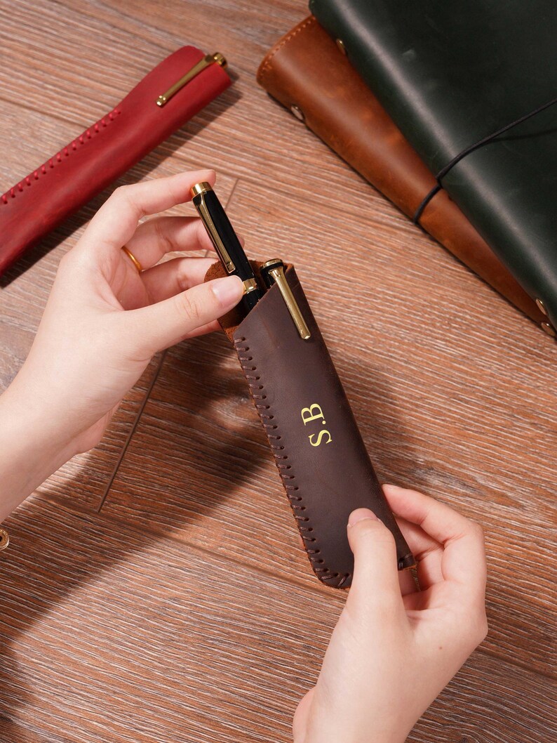 Może przedstawiać: Brązowe sk&oacute;rzane etui na długopis ze złotymi elementami i inicjałami "S.B." w złotych literach. Czarny długopis ze złotymi akcentami jest częściowo włożony. Widoczne są inne sk&oacute;rzane etui w kolorze czerwonym, brązowym i zielonym.