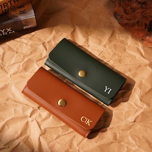 Könnte beinhalten: Zwei Leder-Schlüsseletuis, eines in Waldgrün und das andere in warmem Braun, werden auf einer strukturierten braunen Oberfläche präsentiert. Jedes Schlüsseletui verfügt über einen goldenen Druckknopfverschluss und personalisierte Initialen. Ein stilvolles Accessoire.