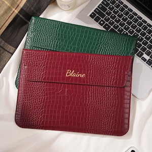 May include: Two laptop sleeves, one green and one burgundy, with a crocodile skin texture. The burgundy sleeve has the name "Blaine" in gold lettering. A laptop and a candle are in the background.