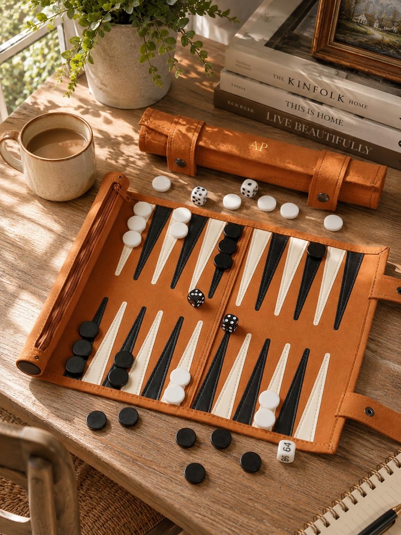 May include: A backgammon set with a tan leather case and board. The board is open, showing black and white game pieces and dice. A mug of coffee and decorative books are also visible.