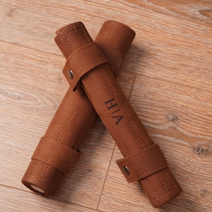 Two rolled-up leather backgammon sets placed on a wooden surface, each featuring personalised initials on the exterior strap. The compact roll-up structure highlights easy storage and travel readiness.