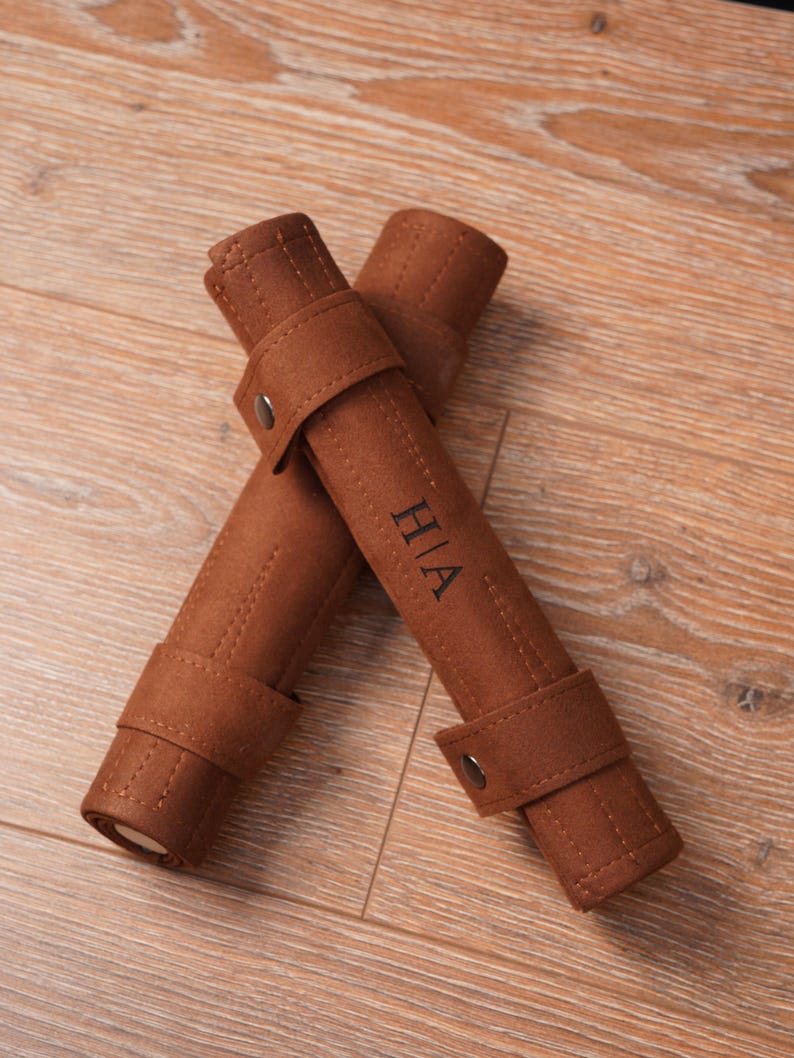 Two rolled-up leather backgammon sets placed on a wooden surface, each featuring personalised initials on the exterior strap. The compact roll-up structure highlights easy storage and travel readiness.