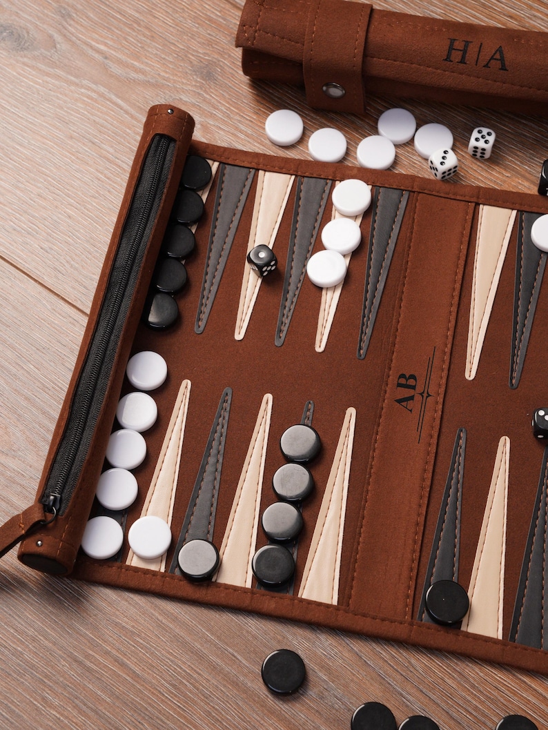 A detailed view of the leather travel backgammon board with black and white checkers in play. Custom initials are engraved on the centre panel, showcasing personalisation options on the roll-up design.