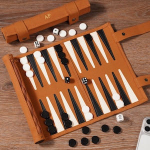 May include: A brown backgammon set with black and white game pieces, dice, and a carrying case. The board is open, displaying the triangular points. The carrying case has the initials "A.P." embossed on it. The game is set on a wooden surface.