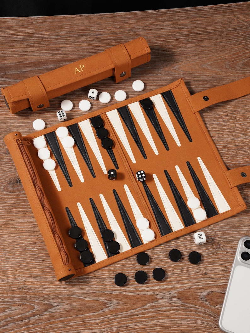 May include: A brown backgammon set with black and white game pieces, dice, and a carrying case. The board is open, displaying the triangular points. The carrying case has the initials "A.P." embossed on it. The game is set on a wooden surface.
