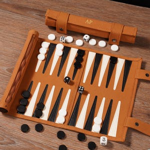 May include: A brown leather backgammon set with white and black game pieces. The set is rolled out on a wooden surface, with dice and a carrying case with the initials "AP".
