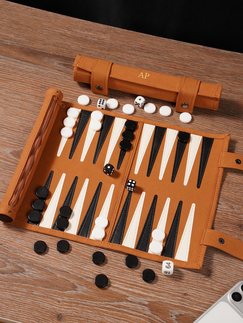 May include: A brown leather backgammon set with white and black game pieces. The set is rolled out on a wooden surface, with dice and a carrying case with the initials "AP".