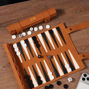 Könnte beinhalten: Ein Backgammon-Set aus braunem Leder mit schwarzen und weißen Spielsteinen, Würfeln und einer passenden Tragetasche. Auf der Tasche sind die Initialen "S|B" eingeprägt. Das Spielbrett ist geöffnet und spielbereit, mit einem Smartphone in der Ecke.