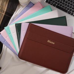 May include: A collection of colorful laptop sleeves in various pastel shades, including pink, mint green, and lavender. The brown sleeve in the foreground is personalized with the name "Blaine" in gold lettering. A laptop and candle are in the background.