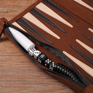 Close-up of the zippered storage pocket on a leather roll-up travel backgammon board, neatly holding black and white checkers and dice. The image highlights the compact, portable design and organized interior ideal for travel use.