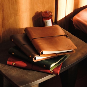 May include: A stack of leather journals in various colors: brown, dark green, and red. The top journal is brown with the initials "SB" embossed. Each journal has a strap closure and brass-colored rivets. The journals are on a dark wooden surface.