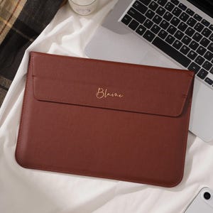 Könnte beinhalten: Eine braune Laptoptasche mit Klappenverschluss. Das Wort "Blaine" ist in goldener Schrift auf der Vorderseite geschrieben. Ein Laptop, eine Kerze und ein Telefon befinden sich im Hintergrund.