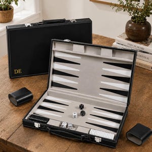 Könnte beinhalten: Ein schwarzes Backgammon-Spiel mit lederartiger Außenseite und den goldenen Initialen DE. Das geöffnete Gehäuse zeigt ein hellgraues Innenleben mit weißen und schwarzen Dreiecksspitzen, schwarzen und weißen Spielsteinen, zwei Würfelbechern und vier Würfeln.