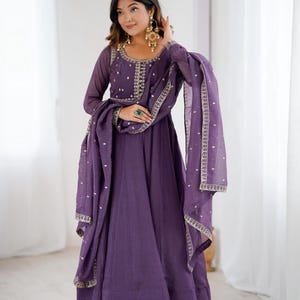 Könnte beinhalten: Ein langes, fließendes lila Kleid mit aufwendiger Stickerei am Ausschnitt und an den Ärmeln. Das Kleid wird mit einem passenden Schal und goldenen Ohrringen kombiniert. Das Outfit ist im traditionellen Stil gehalten und eignet sich für besondere Anlässe.