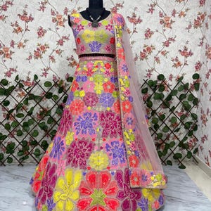 May include: A vibrant, floral-embroidered lehenga choli set. The ensemble features a pink skirt and matching top, adorned with colorful flower patterns in yellow, red, purple, and green. A sheer, pink dupatta with embroidered trim completes the look.