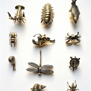 May include: A collection of brass-toned metal figurines of various sea creatures and insects, including a lobster, fish, beetle, anglerfish, crab, snail, dragonfly, turtle, and a sea shell. The figurines are arranged on a white background.