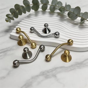 May include: A collection of decorative cabinet hardware in various metallic finishes, including gold, silver, and pewter. The hardware features curved handles with spherical ends and matching round base plates. Eucalyptus sprigs add a natural accent to the display.