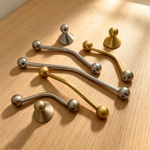 May include: An assortment of metallic cabinet hardware, including curved handles and knob-style pulls. The handles have a ball at each end and are in silver and gold tones. The knobs are round and sit on a circular base. The hardware is arranged on a light wood surface.
