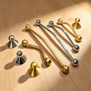 May include: An assortment of cabinet hardware in gold and silver finishes. The collection includes curved pulls with spherical ends and round, conical knobs. The hardware is displayed on a light wood surface.