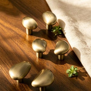 Pode incluir: Uma caneca de cerâmica branca com uma alça preta e o texto "But First, Coffee" em preto, com uma fonte manuscrita. A caneca está sobre uma superfície branca.